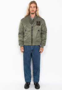 Olijfgroen bomberjack van glanzend nylon, met een ritssluiting aan de voorkant, zijzakken en een zwart stoffen etiket op de linkerkant van de borst.