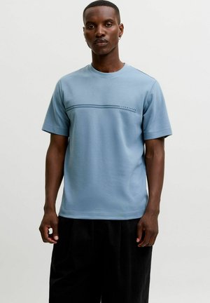 Mand iført en lyseblå t-shirt med korte ærmer med en tynd horisontal stribe og sorte bukser, stående mod en ensfarvet lys baggrund.