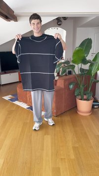 Crna oversized majica s bijelim horizontalnim prugama, kratkim rukavima i okruglim izrezom, drži ju osoba u dnevnoj sobi.