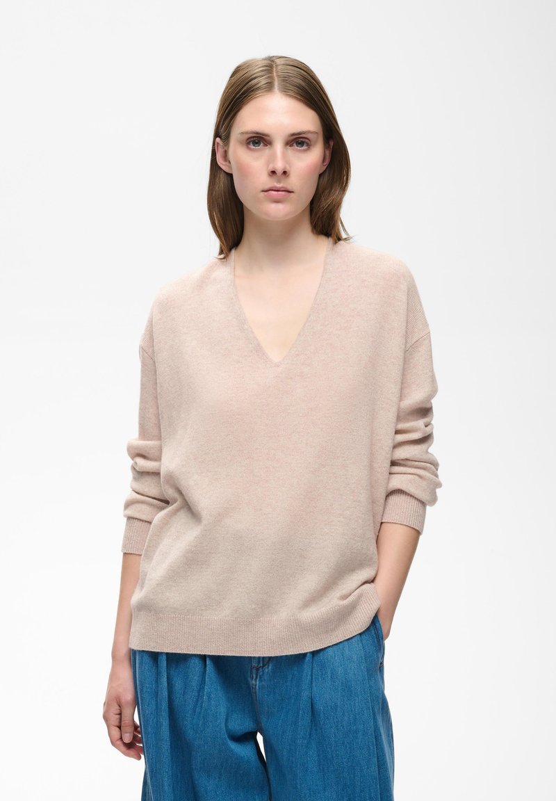 Femme aux cheveux bruns raides, portant un pull beige clair à col en V et un pantalon bleu ample, debout avec une main dans la poche.
