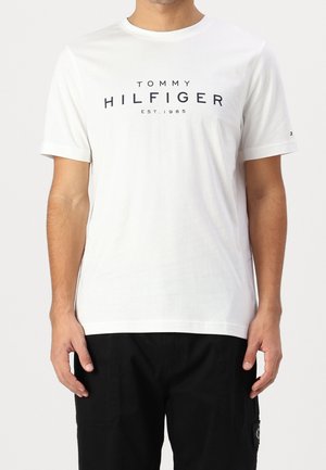 Uomo che indossa una maglietta bianca a maniche corte con la scritta "Tommy Hilfiger EST. 1985", abbinata a pantaloni neri su sfondo semplice.