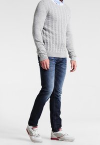 Grå kabelstickad tröja, blå jeans och vita sneakers med röda accenter. Tröjan har en ribbad halsringning och muddar. Jeans har rak passform.