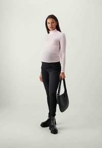 Maglietta a maniche lunghe rosa chiaro, jeans neri aderenti, stivaletti neri con suola spessa e una piccola borsa nera, in piedi su uno sfondo neutro.