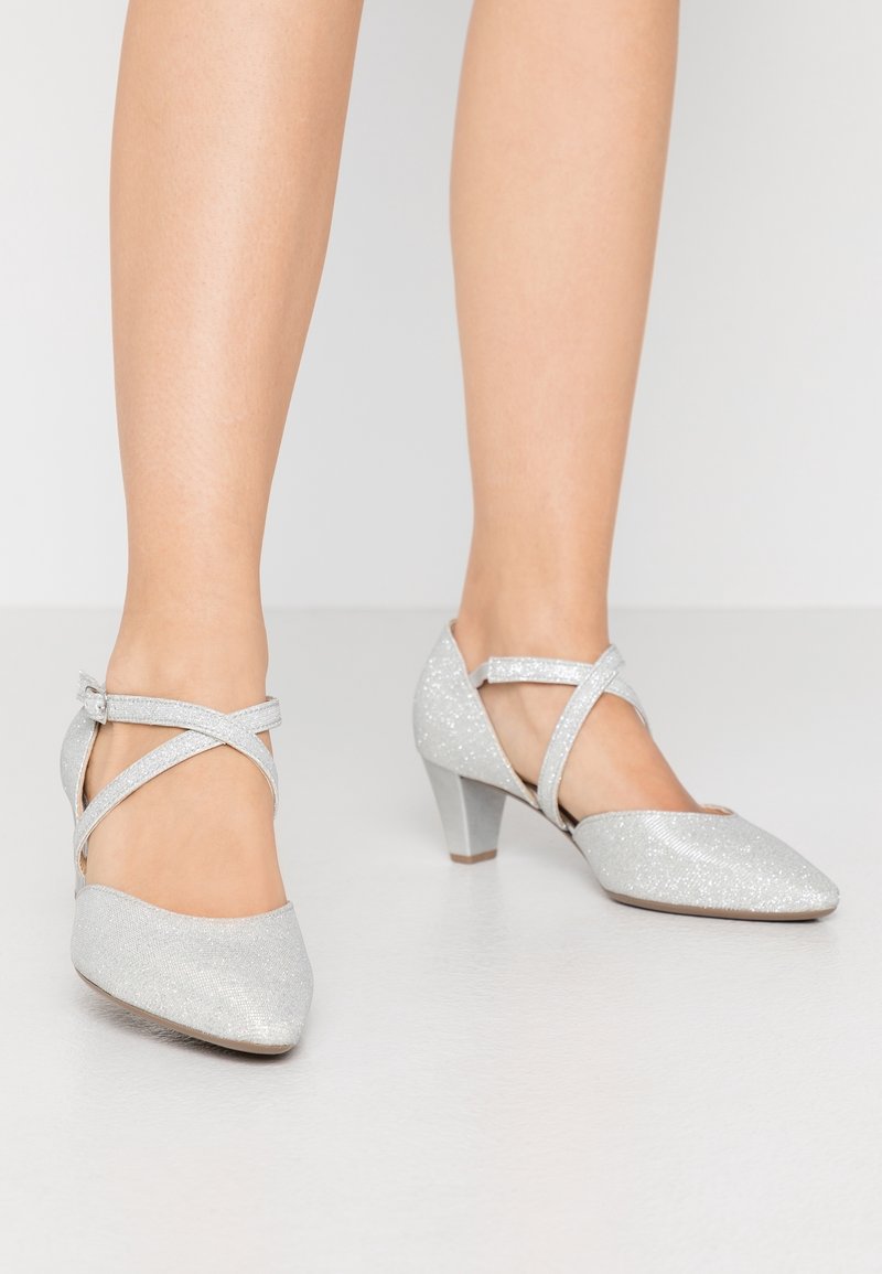 Chaussures à talons hauts pailletées argentées avec un bout pointu, un design à brides croisées à la cheville et un talon effilé, reposant sur une surface claire.