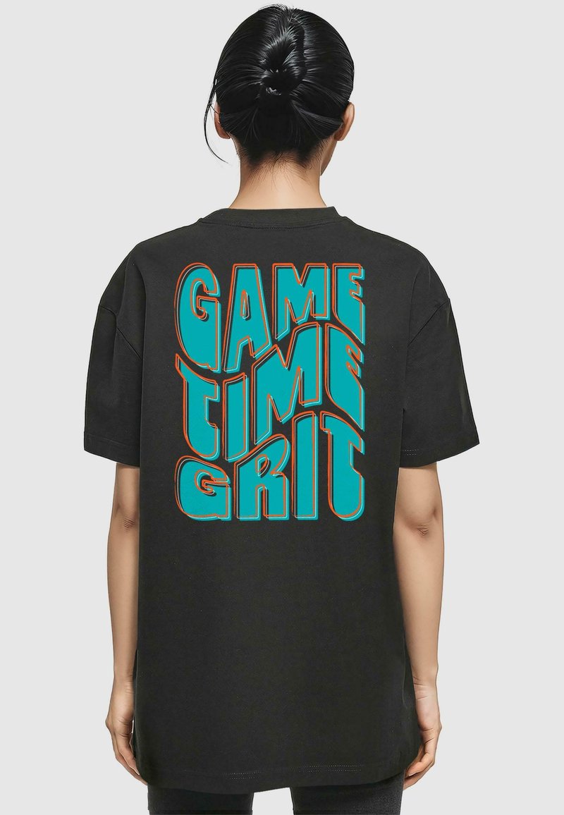 Svart oversized T-skjorte med dristig turkise og oransje bokstaver som viser teksten "GAME TIME GRIT" på baksiden. Myk bomullsmateriale.