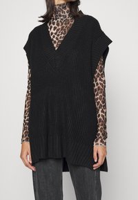 Svart stickad väst med djup V-ringning och vida ärmhål, lager-på-lager över en brun leopardmönstrad långärmad skjorta. Texturerad stickning, avslappnad passform.