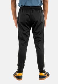 Zwarte sportbroek met een gladde textuur, voorzien van witte zijkanten. Draagt met blauwe en gele sneakers, gezien vanaf de achterkant.