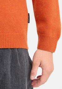 Oransje strikket genser med ribbestrikkede mansjetter og et uformelt merke. Bærerens hånd berører grå denimbukser, noe som viser en kontrast i tekstur.