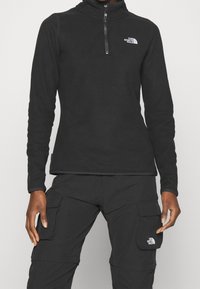 Černý fleecový pulovr s vysokým límcem, čtvrtinovým zipem vpředu a vyšitým logem na hrudi. Hladká textura a slim fit design.