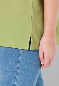 Camicia verde oliva chiaro a maniche corte in tessuto strutturato con spacchi laterali. Abbinata a jeans in denim blu, che mette in evidenza la silhouette del capo.