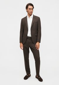 Άνδρας με καφέ κοστούμι και λευκό πουκάμισο, όρθιος με χαλαρά τα χέρια, κοιτάζοντας δεξιά, φοράει σκούρα καφέ μοκασίνια σε απλό ανοιχτό φόντο.