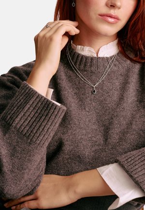 Grauer Strickpullover mit gerippten Bündchen, getragen über einem weißen Hemd. Enthält eine silberne mehrlagige Halskette mit einem schwarzen Anhänger und passenden Ohrringen.