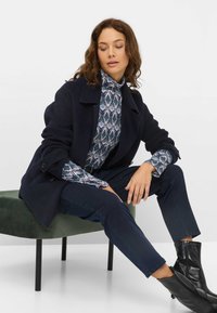 Marine uld frakke med store klapper og knapper over en mønsteret mock turtleneck. Modelen har sat det sammen med mørke jeans og sorte ankelstøvler.