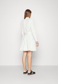 Biała sukienka koszulowa z długimi rękawami, dopasowaną talią i rozkloszowanym dołem; widok z tyłu pokazuje szczegóły guzików oraz gładką teksturę. Uzupełniona o czarne trampki.