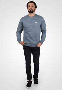 Hanorac din material bleu închis heathered, cu guler rotund și logo triunghiular pe piept, asociat cu blugi negri și adidași negri.