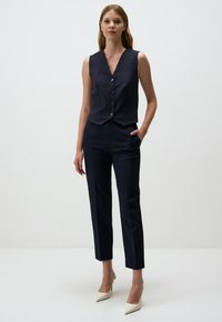 Marineblauwe pinstripe gilet met een v-hals, vijf knopen en bijpassende op maat gemaakte broek. Model draagt crèmekleurige puntige hakken, met handen in de zakken.