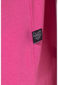 Felroze stof met zichtbare naad en een klein zwart geweven label met witte tekst dat in de zijkant is genaaid.