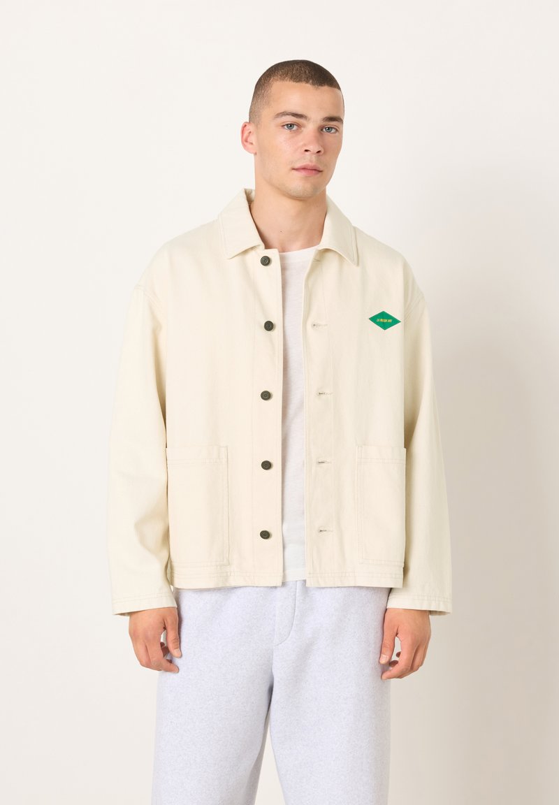 Beige Jeansjacke mit grünem Logo, geradem Schnitt, Knopfleiste, aufgesetzten Taschen und langen Ärmeln. Kombiniert mit grauen Jogginghosen.