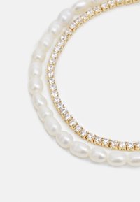 Två halsband: det nedre har oregelbundna vita pärlor, medan det övre har glittrande klara stenar i guldhårdvara.