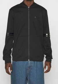 Felpa nera con zip e texture a coste, caratterizzata da due stretti strisce nere su ogni manica e un piccolo logo sul petto. Tasche frontali visibili.