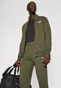 Homme portant un survêtement Puma vert olive et un t-shirt noir, tenant un sac de sport noir avec plusieurs poignées dans la main droite.