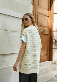 Gilet tricoté crème avec un motif à mailles ouvertes, sans manches, associé à une chemise bleu clair et un pantalon foncé. Porte en bois en arrière-plan.