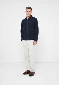 Ναυτικό μπουφάν με φερμουάρ μπροστά και δύο τσέπες στο στήθος, συνδυασμένο με ανοιχτό γκρι παντελόνι και σκούρα καφέ παπούτσια, φοριέται από έναν όρθιο ανδρικό μοντέλο.