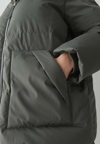 Giacca a vento verde scuro con finitura lucida, dotata di chiusura a zip e ampio tasca laterale con dettaglio a bordo piegato.