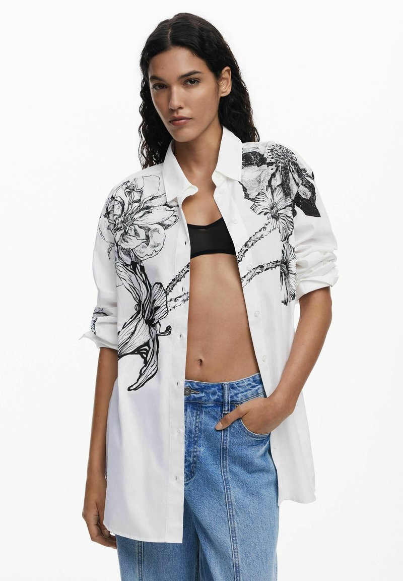 Camicia bianca con bottoni, caratterizzata da disegni floreali neri su una spalla; vestibilità morbida; indossata sopra un top nero; abbinata a jeans in denim azzurro chiaro.
