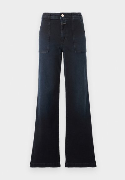 Jean bleu foncé taille haute, jambe large, avec poches plaquées devant, passants pour ceinture et fermeture par bouton sur fond uni clair.