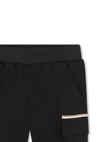 Pantaloni neri con vita a costine, tasche laterali e una tasca decorata con tre strisce orizzontali: oro, bianco e nero. Tessuto testurizzato.