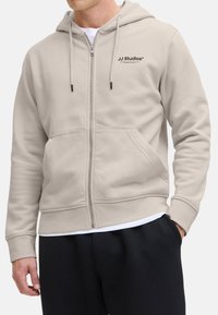 Hellbeige Zip-Hoodie aus weichem Material, mit einer Fronttasche und verstellbarem Kordelzug an der Kapuze sowie einem kleinen Logo auf der Brust.