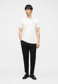 Άνδρας που στέκεται με τα χέρια στις τσέπες, φορώντας λευκό πόλο με κοντά μανίκια και χρυσό έμβλημα, μαύρο παντελόνι και μαύρα επίσημα παπούτσια.