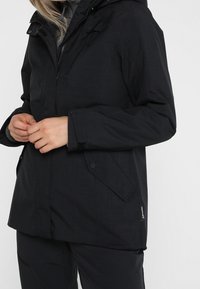 Black waterproof jacket with a hood, featuring a textured fabric, front pockets, and adjustable cuffs. Visible logo on the chest area.