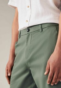 Grüne Chinos mit glatter Textur, ausgestattet mit einem Knopfverschluss, seitlichen Taschen und Gürtelschlaufen, kombiniert mit einem weißen Kurzarmhemd.