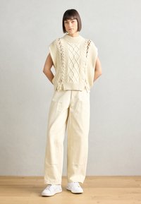 Maglione cable-knit avorio con collo alto, caratterizzato da tagli e dettagli di lacci, abbinato a pantaloni larghi di colore chiaro e sneaker bianche.