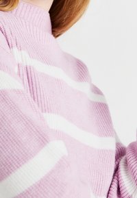 Jasnoróżowy sweter z dzianiny z białymi, poziomymi paskami, z wysokim kołnierzem i prążkowaną fakturą, podkreślający jego przytulny design.