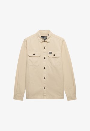 Chemise beige à manches longues avec col, deux poches à rabat sur la poitrine avec boutons, et une petite étiquette de marque au-dessus de la poche droite.