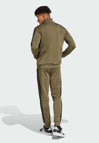 Olivgrön träningsoverall med svarta sidostrmar, jacka med dragkedja och krage, slim fit byxor och svarta sneakers med vita detaljer. Slät, lättviktig tyg.