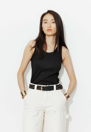 Jeune femme aux longs cheveux foncés porte un haut noir sans manches, un pantalon blanc avec une ceinture noire à clous, et de larges bracelets dorés, debout contre un mur blanc.