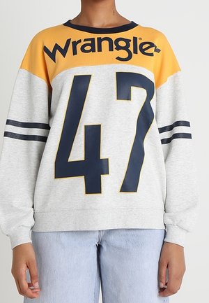 Person trägt einen grauen und gelben Wrangler-Sweatshirt mit marineblauer Zahl 47 und Streifen an den Ärmeln, kombiniert mit hellblauen Jeans.