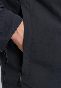 Detail einer Marinejacke mit einer sichtbaren Hand in der Seiten Tasche. Der Stoff ist strukturiert mit einer glatten Oberfläche und zeigt dezente Nähte sowie ein Markenlabel.
