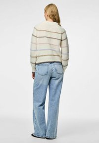 Žena s dugom plavom kosom nosi kremasti pulover sa horizontalnim prugama i široke svijetloplave traperice, stoji naslonjena na jednostavnu pozadinu.