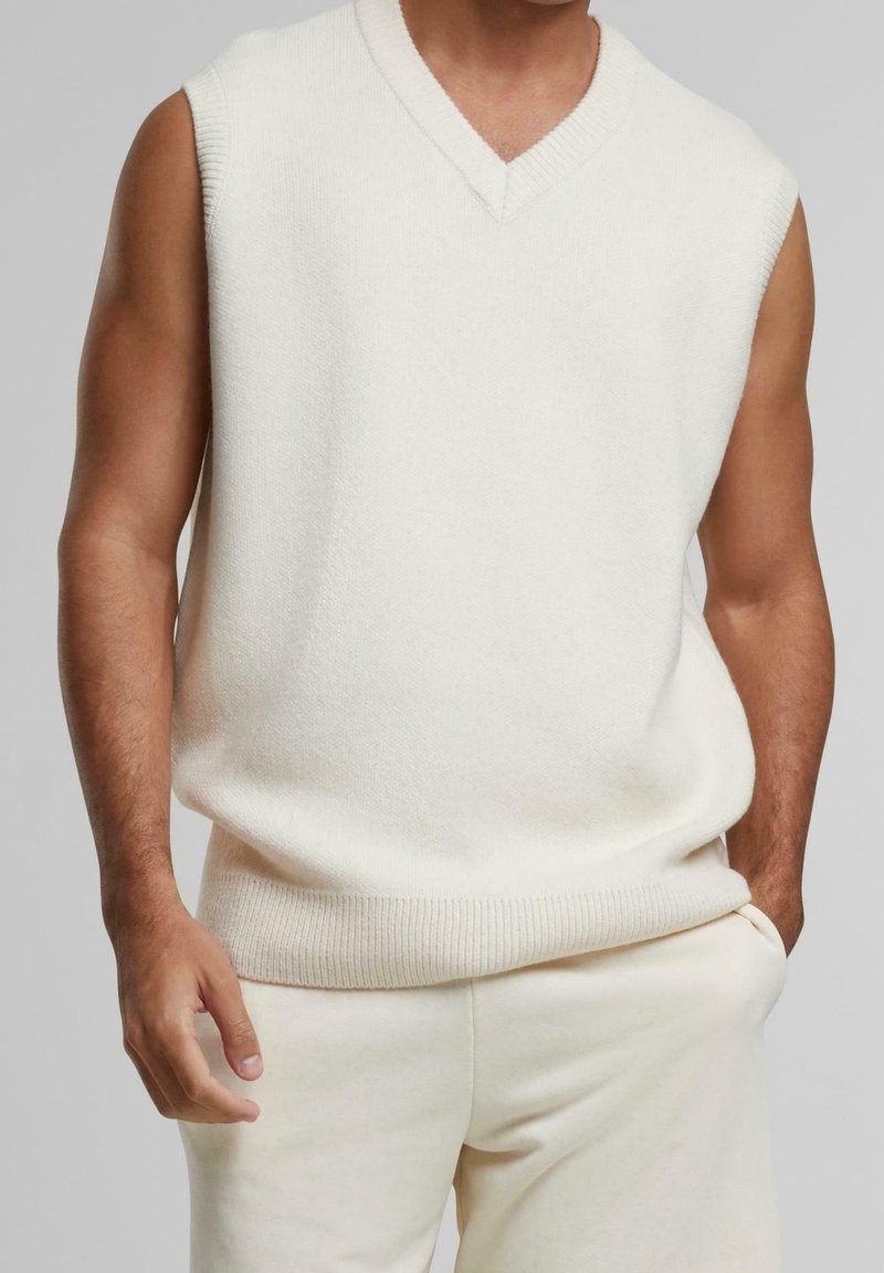 Pull sans manches crème en maille avec un col en V, ourlet côtelé et tissu texturé, associé à un pantalon assorti de couleur claire.
