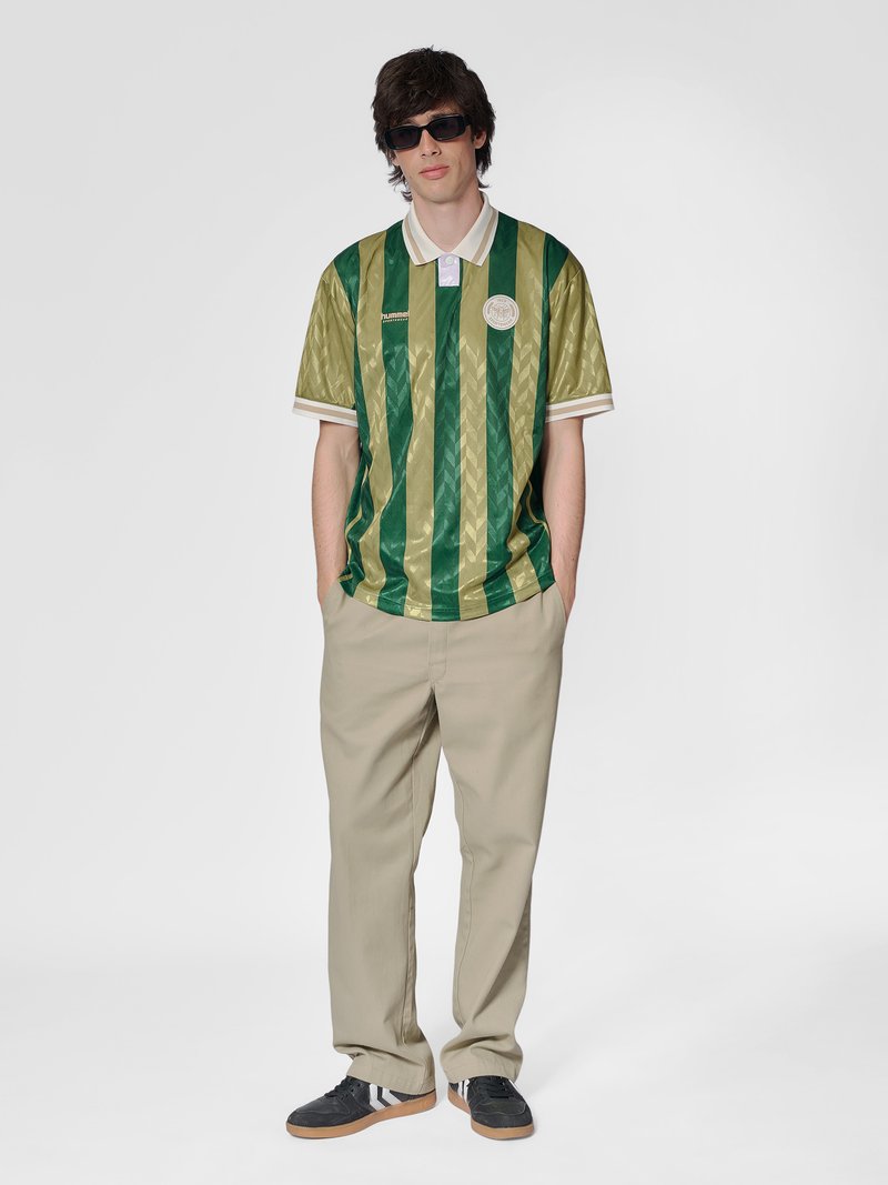 Jeune homme portant des lunettes de soleil foncées, un polo à rayures vertes et or, un pantalon beige et des baskets noires, debout les mains dans les poches.