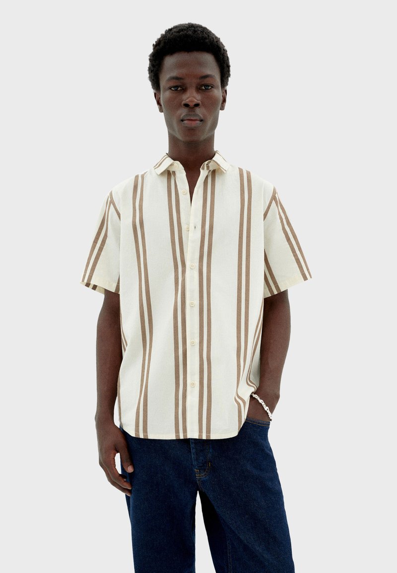 Chemise à col à manches courtes beige avec des rayures verticales marron, présentant un devant à boutons et une coupe décontractée, associée à un pantalon en denim foncé.