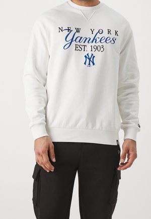 Mężczyzna w białej bluzie z napisem i logo "New York Yankees Est. 1903", w połączeniu z czarnymi spodniami typu cargo.