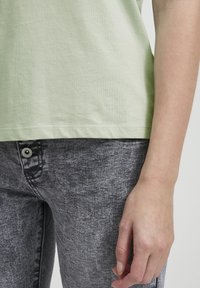 Camiseta de algodón verde claro con dobladillo, combinada con jeans texturizados grises que presentan un botón en la parte delantera y sutiles detalles de costura.