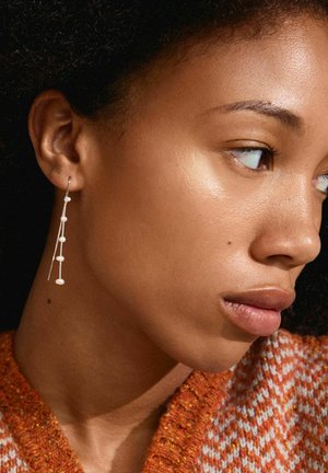Zilveren hangende oorbellen hebben een lineair ontwerp met kleine ronde en ovale kralen. Het model heeft een close-up profielweergave.