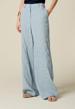 Pantalon large taille haute bleu clair à fines rayures blanches, porté avec un haut noir, un bracelet doré et des sandales plates tressées, main dans la poche.